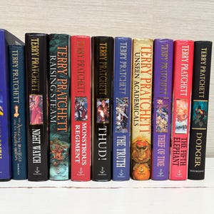 May include: A collection of 11 paperback books by Terry Pratchett, featuring colorful covers with various illustrations and titles including "Going Postal", "Night Watch", "Raising Steam", "Thud!" and "The Fifth Elephant".