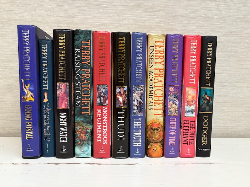 May include: A collection of 11 paperback books by Terry Pratchett, featuring colorful covers with various illustrations and titles including "Going Postal", "Night Watch", "Raising Steam", "Thud!" and "The Fifth Elephant".
