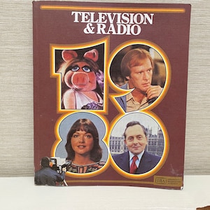 Puede incluir: Una cubierta de libro marrón con el título "Televisión & Radio 1980" en texto blanco. La cubierta presenta cuatro retratos de personas, incluida Miss Piggy de los Muppets.