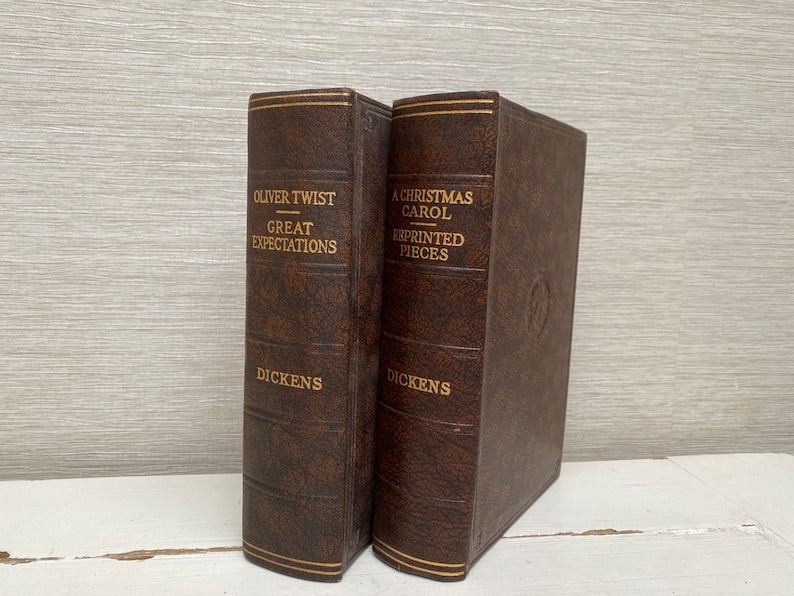 May include: Two antique books with brown leather covers. The titles "Oliver Twist Great Expectations" and "A Christmas Carol Reprinted Pieces" are embossed in gold lettering. The author's name, Dickens, is also in gold. The books have gold trim.