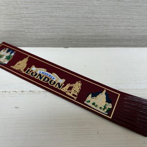 May include: A burgundy bookmark with gold accents and the word "LONDON" in white. The bookmark features illustrations of London landmarks. The bottom edge has a fringed design.