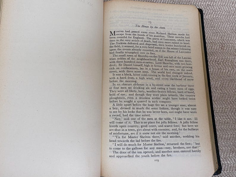 May include: Open book with text on aged paper. The text is in English, with the title "The House by the shore" at the top of the page. The pages are yellowed with age, and the binding is black.