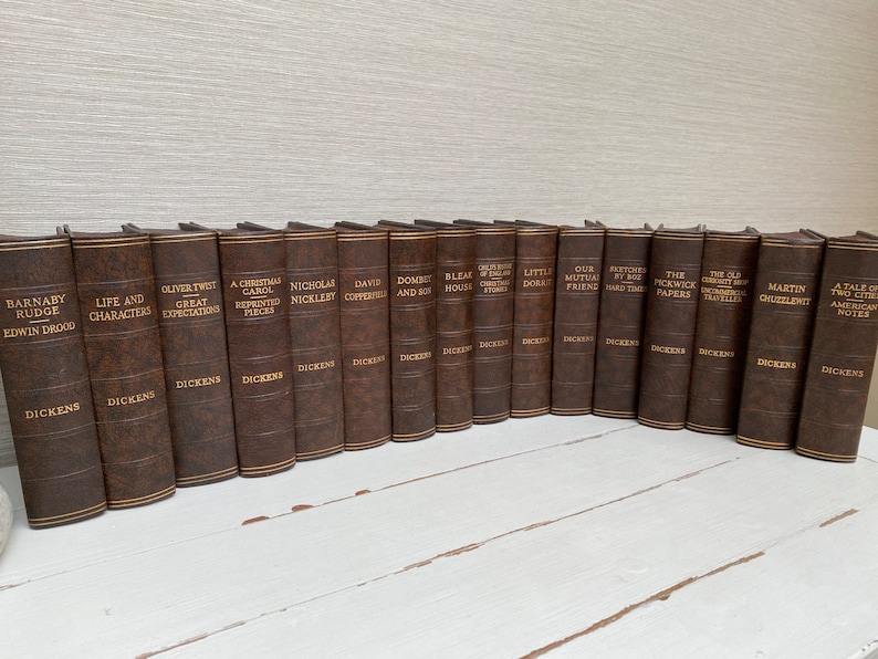 May include: A collection of brown leather-bound books with gold lettering, featuring titles by Charles Dickens. The spines display titles like "Oliver Twist," "A Christmas Carol," and "David Copperfield." The books are arranged on a white surface.