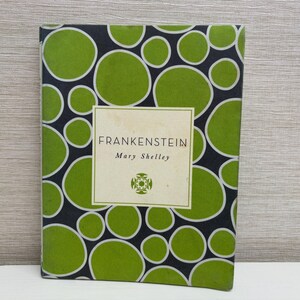 Könnte beinhalten: Ein Buchcover mit schwarzem Hintergrund und grünen Kreisen mit weißen Umrissen. Der Titel "Frankenstein" und der Autor "Mary Shelley" sind in Weiß auf einem weißen Rechteck in der Mitte des Covers gedruckt.