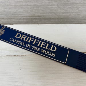 May include: A navy blue bookmark with the text "DRIFFIELD CAPITAL OF THE WOLDS" in a serif font. The bookmark has a decorative crest at the top left and a series of vertical lines at the bottom.