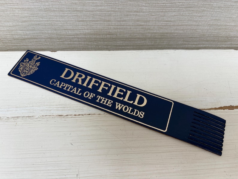 May include: A navy blue bookmark with the text "DRIFFIELD CAPITAL OF THE WOLDS" in a serif font. The bookmark has a decorative crest at the top left and a series of vertical lines at the bottom.