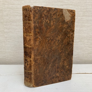 Puede incluir: Un libro antiguo encuadernado en cuero marrón con detalles dorados y una cubierta de mármol. El libro está ligeramente desgastado y tiene una etiqueta descolorida en el lomo.