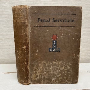 以下が含まれることがあります： 表紙に「Penal Servitude」というタイトルが書かれた茶色の本。表紙には赤い星と、「5 Y 1 3 1」という文字が書かれたブロックの積み重ねも描かれている。