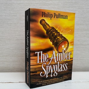 Peut inclure: Un livre relié intitulé "The Amber Spyglass" de Philip Pullman. La couverture présente une longue-vue dorée sur fond de coucher de soleil flamboyant. La tranche est noire avec le titre et le nom de l'auteur.