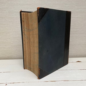 May include: A vintage hardcover book with a dark blue cover and aged pages. The book is closed and standing upright on a white surface.