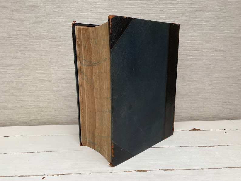 May include: A vintage hardcover book with a dark blue cover and aged pages. The book is closed and standing upright on a white surface.