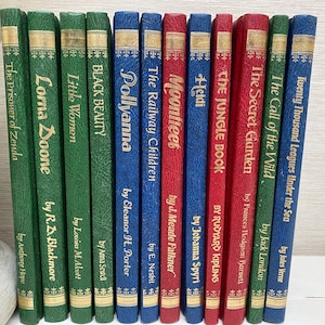 Puede incluir: Una colección de 14 libros antiguos de tapa dura con letras doradas en los lomos. Los libros están dispuestos verticalmente y son principalmente verdes, azules y rojos. Los títulos incluyen "El prisionero de Zenda", "Lorna Doone", "Mujercitas", "Black Beauty", "Pollyanna", "Los niños del ferrocarril", "Moonfleet", "Heidi", "El libro de la selva", "El jardín secreto", "La llamada de la selva", "Veinte mil leguas de viaje submarino".