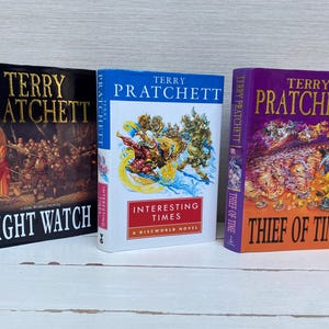 May include: Three hardcover books by Terry Pratchett, featuring colorful covers with illustrations. The titles are "Night Watch", "Interesting Times", and "Thief of Time".