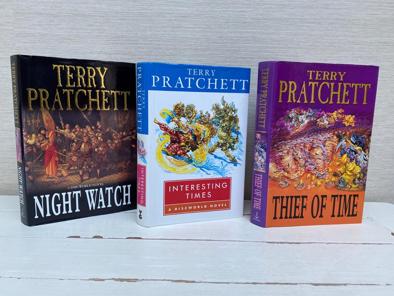 May include: Three hardcover books by Terry Pratchett, featuring colorful covers with illustrations. The titles are "Night Watch", "Interesting Times", and "Thief of Time".