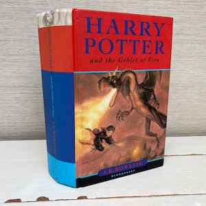 Puede incluir: Un libro de tapa dura rojo con una columna vertebral azul. La portada presenta una ilustración de un dragón que escupe fuego y Harry Potter volando en una escoba. El título es "Harry Potter y el Cáliz de Fuego" de J.K. Rowling.