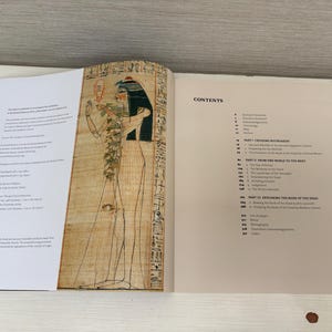 May include: An open book with a black spine, displaying pages with text and an illustration of an Egyptian figure. The right page features a "CONTENTS" section. The illustration is of a figure with a black headdress and hieroglyphs.