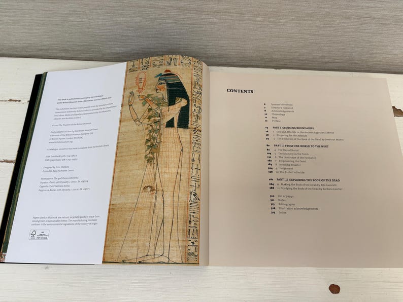 May include: An open book with a black spine, displaying pages with text and an illustration of an Egyptian figure. The right page features a "CONTENTS" section. The illustration is of a figure with a black headdress and hieroglyphs.