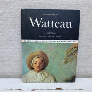 May include: A book titled "L'opera completo di Watteau" by Giovanni Macchia. The book cover features a painting of a man wearing a white hat and a white shirt with a ruffled collar. The background is a blue sky with a green tree.
