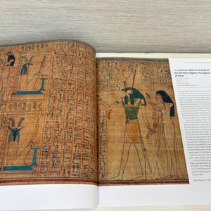 May include: Open book featuring ancient Egyptian art. The left page displays hieroglyphs and figures with blue and black accents. The right page shows a detailed illustration of a figure with a green and gold collar, accompanied by text titled "A woman's book of the Dead of the late New Kingdom: the papyrus of Anhai."