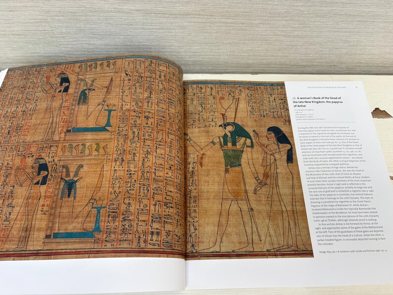 May include: Open book featuring ancient Egyptian art. The left page displays hieroglyphs and figures with blue and black accents. The right page shows a detailed illustration of a figure with a green and gold collar, accompanied by text titled "A woman's book of the Dead of the late New Kingdom: the papyrus of Anhai."