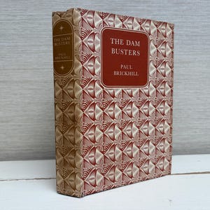May include: A vintage hardcover book, "The Dam Busters" by Paul Brickhill. The cover features a repeating geometric design in red and beige. The title is displayed in a red rectangular frame.