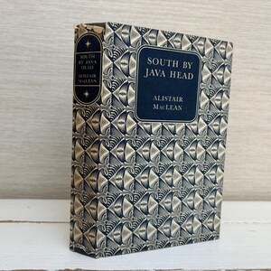 Puede incluir: Un libro antiguo de tapa dura titulado "South by Java Head" de Alistair MacLean. El libro tiene un patrón geométrico azul marino y crema en la portada y el lomo. El título y el nombre del autor están en texto blanco sobre un fondo azul marino.