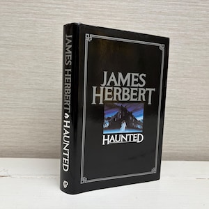 以下が含まれることがあります： ジェームズ・ハーバート著「Haunted」というタイトルの黒いハードカバーの本。表紙には、建物の暗い雰囲気のある画像が描かれており、ドアの入り口に人物が立っている。