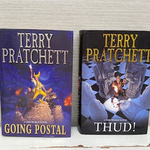 May include: Two paperback books by Terry Pratchett, a Discworld novel titled "Going Postal" and a Discworld novel titled "Thud!"