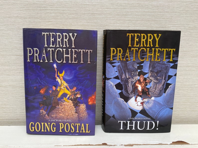 May include: Two paperback books by Terry Pratchett, a Discworld novel titled "Going Postal" and a Discworld novel titled "Thud!"