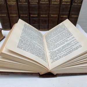 May include: An open book with aged, yellowed pages, bound in a brown leather cover. The book is in front of a row of similar books with titles like "Pickwick Papers" and "Hard Times" by Dickens. The pages are filled with printed text.