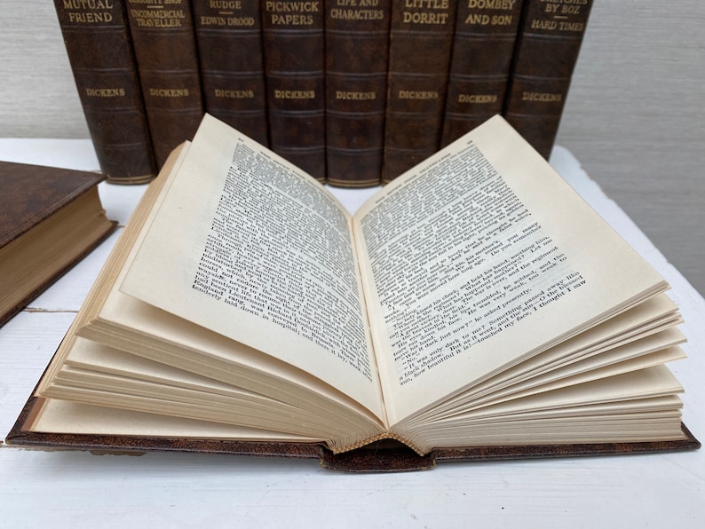 May include: An open book with aged, yellowed pages, bound in a brown leather cover. The book is in front of a row of similar books with titles like "Pickwick Papers" and "Hard Times" by Dickens. The pages are filled with printed text.