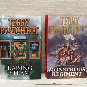 May include: Two paperback books by Terry Pratchett, "Raising Steam" and "Monstrous Regiment", both featuring illustrations and the Discworld logo.