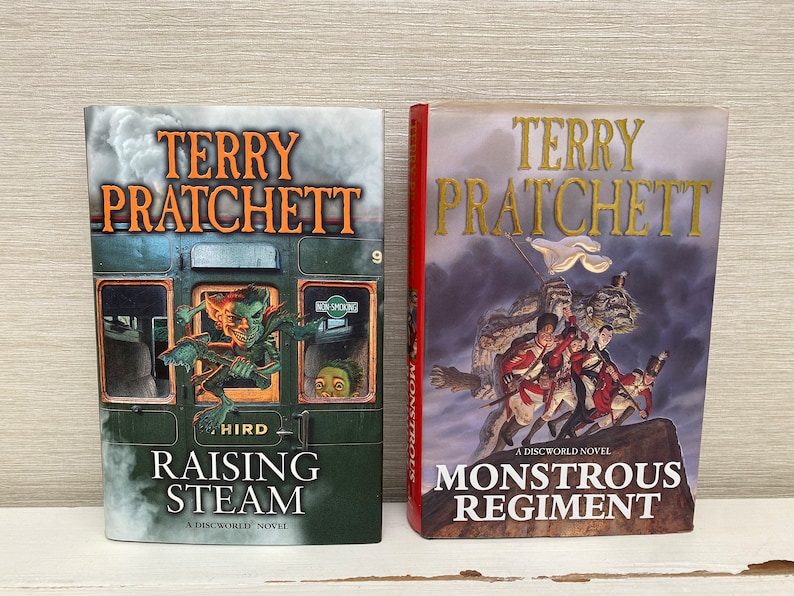 May include: Two paperback books by Terry Pratchett, "Raising Steam" and "Monstrous Regiment", both featuring illustrations and the Discworld logo.