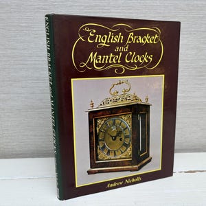 Puede incluir: Un libro marrón titulado "English Bracket and Mantel Clocks" con una esfera de reloj dorada en la portada. El libro es de Andrew Nicholls.