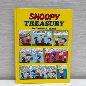 May include: A hardcover book titled "Snoopy Treasury" by Charles M. Schulz. The cover is yellow with the title in red and blue. The book features comic strips with Snoopy and other Peanuts characters.