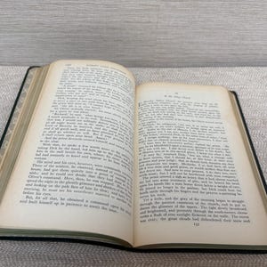 May include: An open book with aged, yellowed pages, revealing printed text. The book has a black cover and is lying on a patterned surface. The text is in English. The book is likely a vintage novel or historical text.