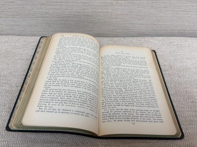 May include: An open book with aged, yellowed pages, revealing printed text. The book has a black cover and is lying on a patterned surface. The text is in English. The book is likely a vintage novel or historical text.