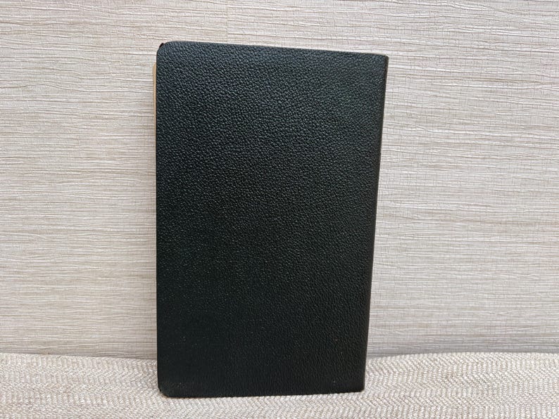 May include: A black, rectangular object with a textured surface. The object is upright and appears to be made of leather or a similar material. The edges are slightly rounded. The background is a light wood grain.