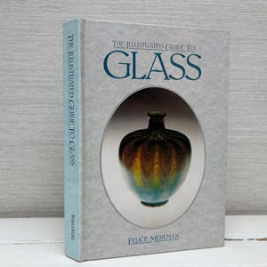 Könnte beinhalten: Der Einband des Buches "The Illustrated Guide to Glass" zeigt eine blaue und grüne Glasvase mit einem wirbelnden Muster. Der Buchtitel ist in weißer Schrift auf grauem Hintergrund.