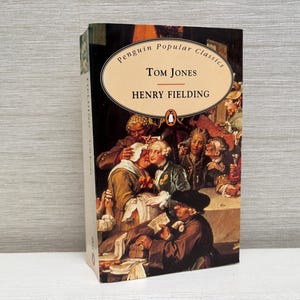 Puede incluir: Una edición Penguin Popular Classics de "Tom Jones" de Henry Fielding. La portada del libro presenta una ilustración detallada de una reunión de personas con ropa de época. El título y el nombre del autor se muestran en forma ovalada en la parte superior.