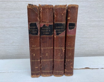 Könnte beinhalten: Vier antike Bücher mit abgenutzten braunen Lederdeckeln. Die Buchrücken zeigen den Titel "SWIFT'S SELECT WORKS" auf schwarzen Etiketten. Die Bücher sind mit 1, 2, 3 und 5 nummeriert. Die Bücher sind vertikal angeordnet.