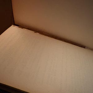 May include: An open book with off-white pages and a light brown cover. The pages are slightly textured, and the binding is visible along the spine. The lighting is soft, creating subtle shadows.