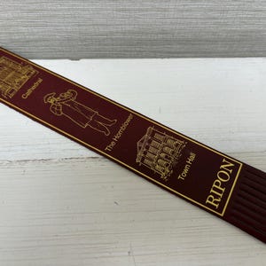 May include: A burgundy bookmark with gold lettering and illustrations. The bookmark features images of a cathedral, a hornblower, and Town Hall, with the word "RIPON" at the bottom. The bookmark has a fringed edge.