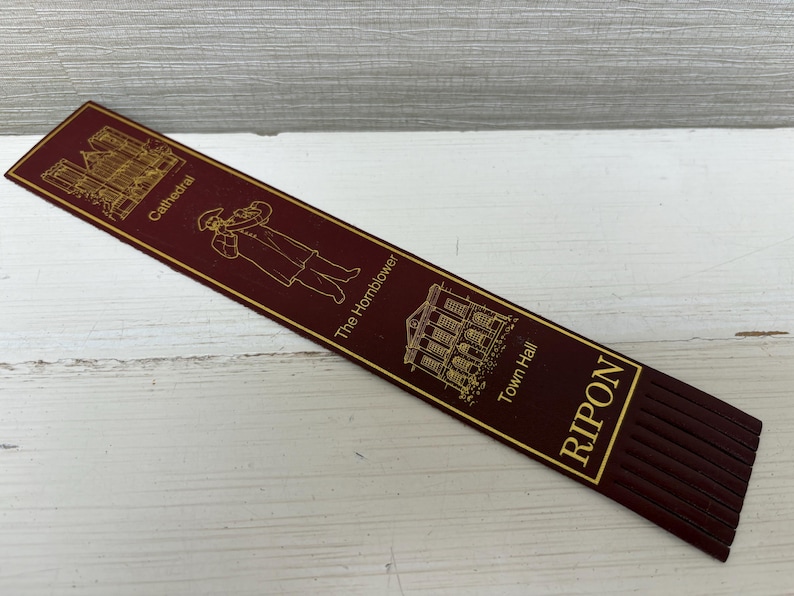 May include: A burgundy bookmark with gold lettering and illustrations. The bookmark features images of a cathedral, a hornblower, and Town Hall, with the word "RIPON" at the bottom. The bookmark has a fringed edge.