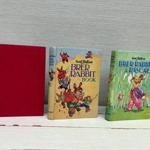 May include: Three vintage children's books are displayed. The books include a solid red book, a book titled "Brer Rabbit Book" with illustrations of rabbits, and a green book titled "Brer Rabbits A Rascal".