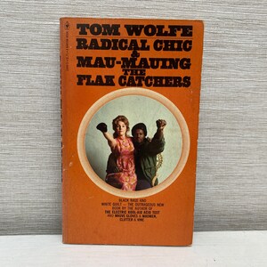 May include: Vintage paperback book titled "Radical Chic & Mau-Mauing the Flak Catchers" by Tom Wolfe. The cover is orange with a circular image of a man and woman with raised fists. The book's title and author are in black text.