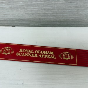 May include: A red rectangular sign with gold lettering and a rose emblem. The text reads "ROYAL OLDHAM SCANNER APPEAL". The sign is on a white surface.