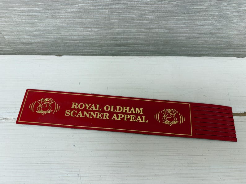 May include: A red rectangular sign with gold lettering and a rose emblem. The text reads "ROYAL OLDHAM SCANNER APPEAL". The sign is on a white surface.