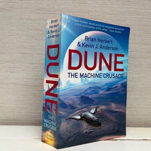 May include: A hardcover book titled "Dune: The Machine Crusade" by Brian Herbert & Kevin J. Anderson. The cover features a landscape with a flying object and the title in large red letters. The spine reads "DUNE".