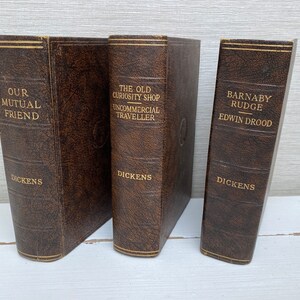 May include: Three antique books with brown leather covers. The titles "Our Mutual Friend", "The Old Curiosity Shop", and "Barnaby Rudge" are printed in gold. The author, Dickens, is also printed on the spines.